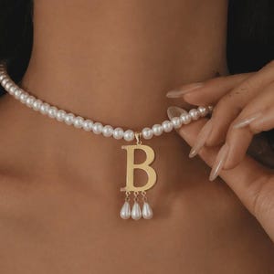 以下が含まれることがあります： 「B」の文字と3つの垂れ下がった真珠のドロップが付いた、金のペンダントが付いた真珠のネックレス。ネックレスは、小さくて丸い白い真珠で構成されています。ペンダントは光沢のある金色です。