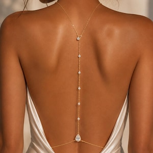 May include: A gold-toned back necklace featuring a delicate chain that drapes down the spine, accented with multiple small round clear stones and a single pear-shaped clear stone at the base, worn with a sleeveless satin garment.