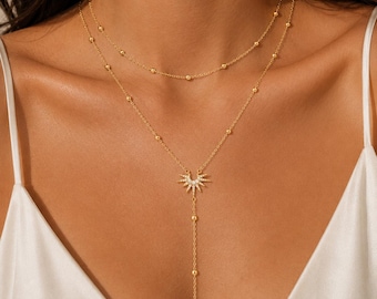 May include: A gold-toned layered necklace featuring two delicate chains. The shorter chain has small evenly spaced beads, while the longer chain includes a sunburst pendant set with clear stones and a vertical drop chain with additional beads.