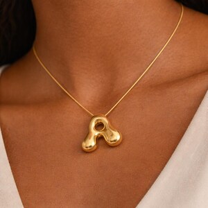 May include: A gold pendant necklace featuring an abstract design. The pendant has a rounded shape with a central hole, suspended from a fine gold chain. The necklace is worn against the skin, creating a striking contrast.