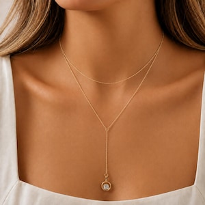 May include: A gold-toned layered necklace featuring a delicate chain that circles the neck and a second, longer chain that drops down to a circular pendant. The pendant contains a single, clear, round-cut stone set in the centre of a gold ring.