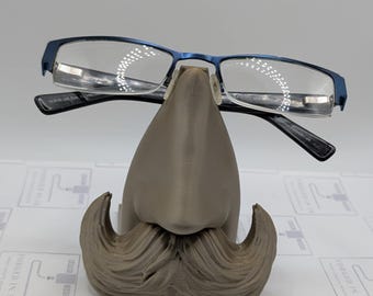 May include: A 3D printed gray mustache and nose stand for holding eyeglasses. The stand has a mustache and a nose with a pair of eyeglasses resting on the nose.