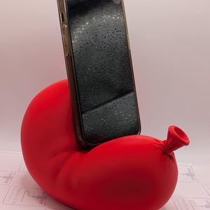 May include: A red, inflatable phone stand shaped like a balloon with a small nozzle. A black smartphone is propped up in the stand.
