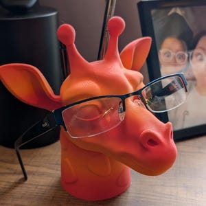 Puede incluir: Un soporte para gafas con forma de jirafa de color rojo y naranja con gafas negras apoyadas en el soporte. El soporte tiene una superficie texturizada y una pequeña nariz redonda.