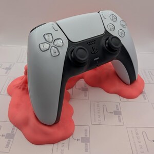 Puede incluir: Un mando de PlayStation 5 blanco y negro descansa sobre un soporte de impresión 3D de color rosa brillante. El soporte tiene un efecto derretido y goteante. El texto "FORGED IN 3D" es visible en la superficie debajo del soporte.