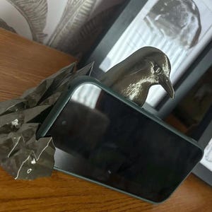 May include: A dark green smartphone case with a bird sculpture emerging from the top. The bird is dark grey and has a long beak. The case is resting on a wooden surface, with framed artwork in the background.