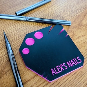 Pode incluir: Uma paleta preta para nail art com detalhes em rosa e o texto "ALEX'S NAILS" em rosa. Duas ferramentas de nail art prateadas também são visíveis. A paleta tem três círculos rosa e ranhuras com bordas rosa.