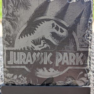 Könnte beinhalten: Ein schwarzer Schieferuntersetzer mit einem grauen Jurassic Park-Logo, das eine Dinosauriersilhouette zeigt. Das Logo ist von einem Blattwerk-Dschungeldesign umgeben.