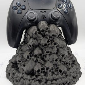May include: A black PlayStation 5 controller sits on a black skull-shaped stand. The stand is made of many small skulls stacked on top of each other.