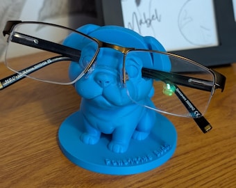 May include: Eyeglasses with a black and gold frame are displayed on a vibrant blue pug-shaped glasses holder. The holder sits on a wooden surface. A framed dog picture is visible in the background.