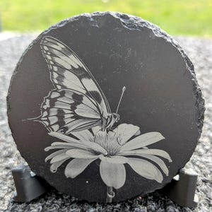 May include: A black slate coaster with a laser-etched image of a butterfly on a flower. The coaster has a black stand.