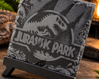Puede incluir: Un posavasos cuadrado de pizarra negra con el logotipo de Jurassic Park grabado, la silueta de un esqueleto de T-Rex y detalles de follaje. El posavasos tiene un borde de piedra natural rugosa y se apoya en un soporte de plástico negro.