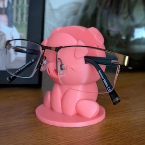 Puede incluir: Un soporte para gafas en forma de cerdo rosa con un par de gafas apoyadas en él. Las gafas tienen una montura dorada y lentes transparentes. El soporte es de plástico y tiene un acabado liso.