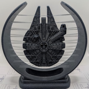 Może przedstawiać: Czarny model statku kosmicznego Millennium Falcon wydrukowany w 3D, zawieszony w czarnej, okrągłej ramie. Rama jest podtrzymywana przez czarną podstawę.
