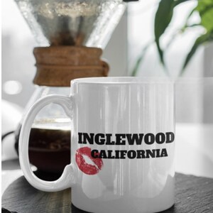 Może przedstawiać: Biały kubek ceramiczny z czarno-białym nadrukiem z napisem "INGLEWOOD CALIFORNIA" i czerwonym śladem po szmince.