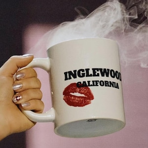 Może przedstawiać: Biały kubek ceramiczny z czarnym napisem "INGLEWOOD CALIFORNIA" i czerwonym odciskiem szminki. Para unosi się z kubka.