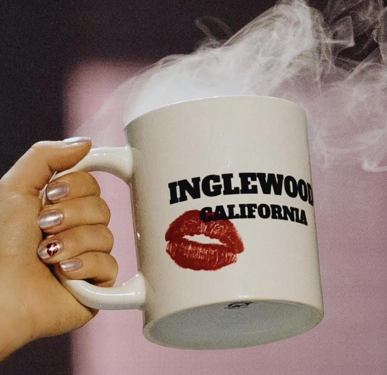 Może przedstawiać: Biały kubek ceramiczny z czarnym napisem "INGLEWOOD CALIFORNIA" i czerwonym odciskiem szminki. Para unosi się z kubka.