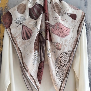 May include: A square silk scarf featuring a cream background with a detailed print of various hot air balloons, trunks, and luggage cases in shades of brown, pink, and black. The scarf has a dark brown border and is draped over a plain white shirt.