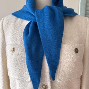 May include: A bright blue knit scarf tied around the neck, worn over a cream-coloured textured jacket featuring two chest pockets and three gold-toned metal buttons with embossed detailing on the front closure and pocket flaps.