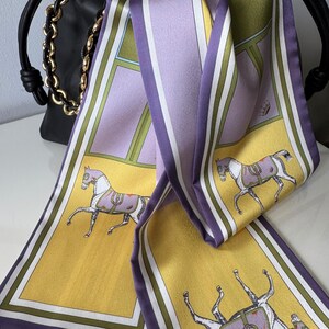 May include: A long rectangular silk scarf featuring a repeating pattern of horses in purple blankets against a yellow background, framed by lavender and white stripes. The scarf is draped over a black leather handbag with a gold chain strap.