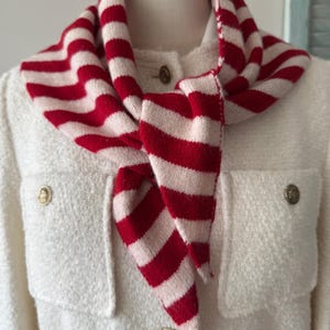 Puede incluir: Una bufanda de punto con rayas horizontales rojas y blanco hueso, atada en un nudo alrededor del cuello de un maniquí que viste una chaqueta texturizada de color blanco hueso con botones dorados y dos bolsillos en el pecho.