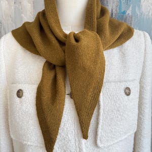 May include: A mustard yellow triangular knit scarf tied around the neck, worn over a white textured jacket featuring two chest pockets and round metallic buttons with an embossed design on the front closure and pocket flaps.