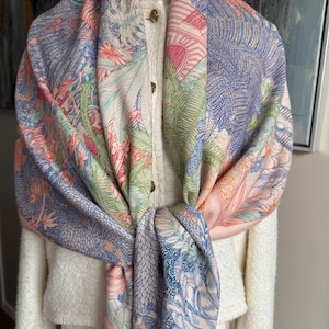 May include: A large square scarf featuring a detailed botanical and architectural print in shades of blue, coral, green, and cream. The scarf is draped over a white textured cardigan with gold buttons, displaying intricate line work of foliage and structures.