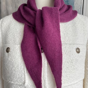 Puede incluir: Una bufanda de punto color magenta intenso con un extremo puntiagudo, colocada alrededor del cuello de un maniquí que viste una chaqueta blanca texturizada con dos bolsillos en el pecho y botones metálicos. La bufanda es suave y esponjosa.