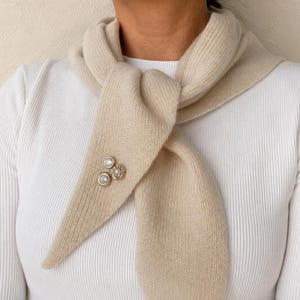May include: A beige knit scarf featuring a ribbed texture, tied around the neck and secured with a silver-toned brooch containing three circular, clear, faceted stones arranged in a cluster on the front end of the fabric.