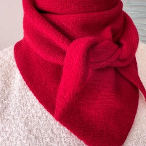 May include: A vibrant red knitted triangular scarf wrapped around a mannequin neck. The scarf features a textured knit pattern and is tied in a simple knot at the front, creating a layered appearance with pointed ends hanging down the chest area.