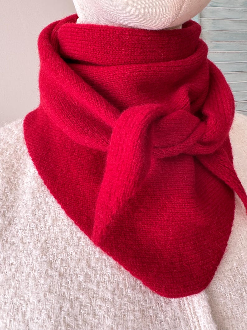 May include: A vibrant red knitted triangular scarf wrapped around a mannequin neck. The scarf features a textured knit pattern and is tied in a simple knot at the front, creating a layered appearance with pointed ends hanging down the chest area.