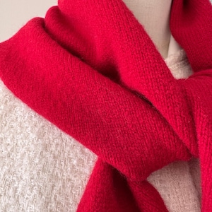May include: A vibrant red knitted scarf draped around the neck, featuring a soft, textured wool-like material. The scarf is layered over a cream-colored, textured tweed-style garment, creating a clear contrast between the bright red and neutral tones.