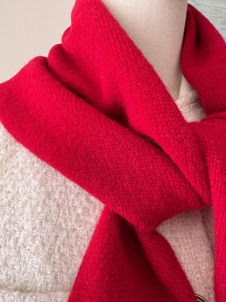 May include: A vibrant red knitted scarf draped around the neck, featuring a soft, textured wool-like material. The scarf is layered over a cream-colored, textured tweed-style garment, creating a clear contrast between the bright red and neutral tones.