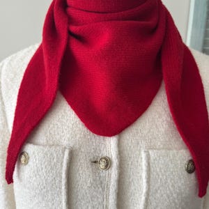 May include: A bright red triangular knit scarf wrapped around the neck, worn over a cream-colored textured jacket featuring two front pockets and three gold-toned metal buttons with embossed circular detailing on the front placket.