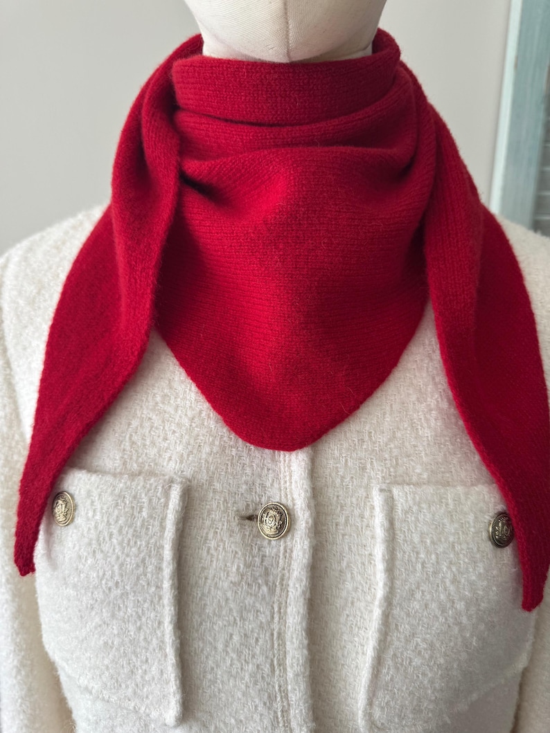May include: A bright red triangular knit scarf wrapped around the neck, worn over a cream-colored textured jacket featuring two front pockets and three gold-toned metal buttons with embossed circular detailing on the front placket.