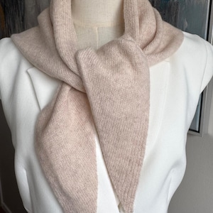 May include: A soft, light beige knit scarf with a triangular shape, draped around the neck of a mannequin wearing a crisp white blazer. The scarf features a fine, textured knit pattern and pointed ends that hang down in front of the chest.