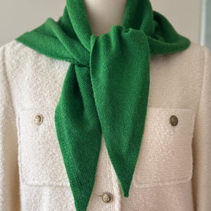 May include: A vibrant green knitted triangular scarf draped around the neck, worn over a cream-coloured textured jacket featuring two front pockets and three metallic buttons with embossed circular designs on the front closure and pocket flaps.
