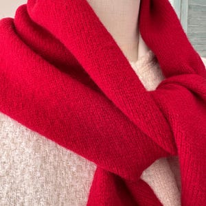 May include: A vibrant red knitted scarf draped around the neck of a mannequin wearing a textured cream-colored garment. The scarf features a soft, fuzzy texture and is wrapped loosely, showing the dense knit pattern of the fabric clearly.