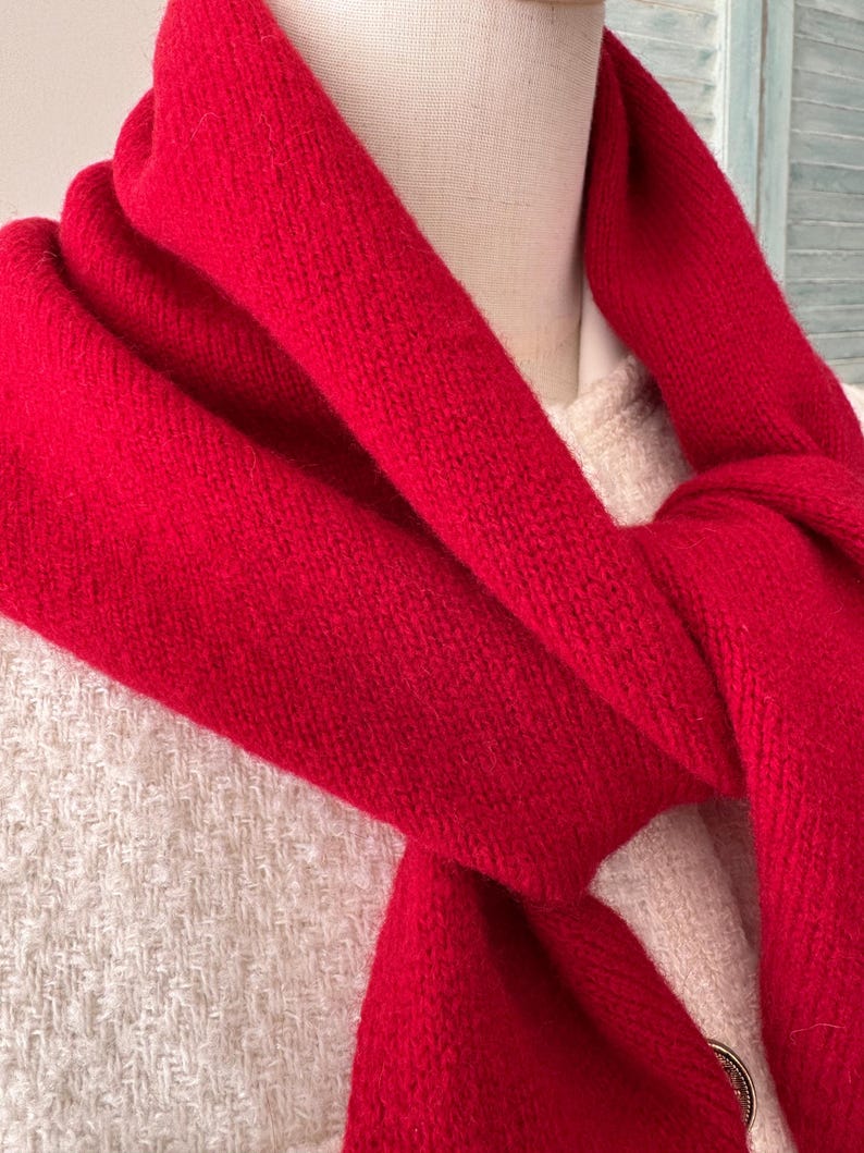 May include: A vibrant red knitted scarf draped around the neck of a mannequin wearing a textured cream-colored garment. The scarf features a soft, fuzzy texture and is wrapped loosely, showing the dense knit pattern of the fabric clearly.