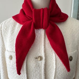 May include: A bright red knitted triangular scarf tied in a knot around the neck, worn over a cream-colored textured jacket featuring two front pockets and three metallic buttons with embossed circular designs on the front placket.