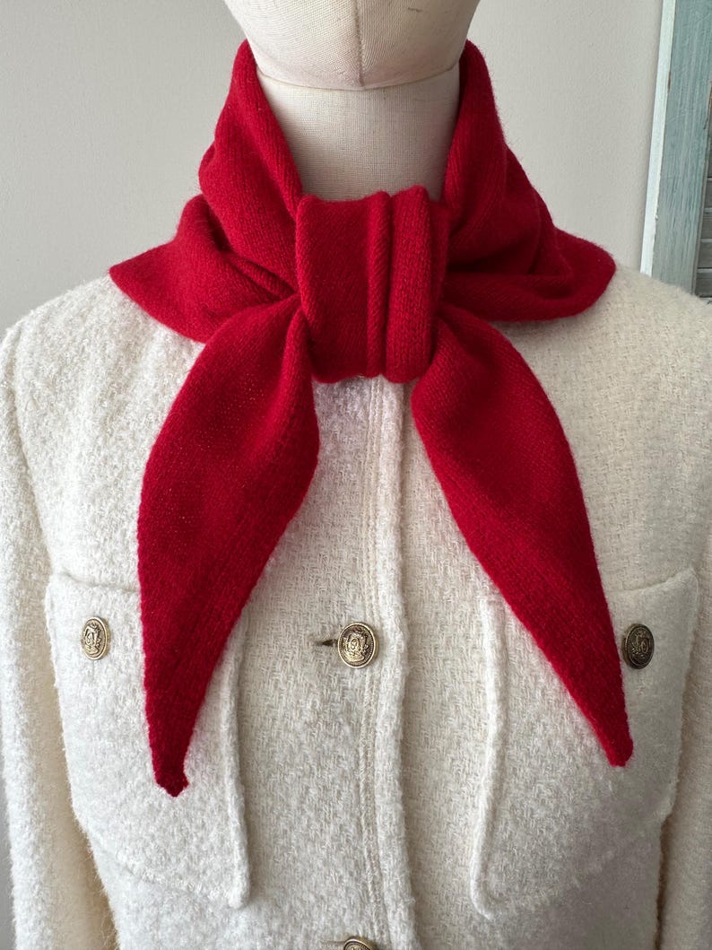 May include: A bright red knitted triangular scarf tied in a knot around the neck, worn over a cream-colored textured jacket featuring two front pockets and three metallic buttons with embossed circular designs on the front placket.