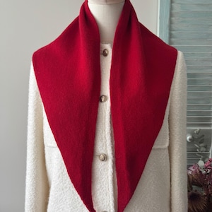 May include: A bright red triangular knit scarf draped over a cream-colored textured jacket. The jacket features a front button closure with three visible metallic buttons. The scarf has a smooth, solid-colored knit texture and pointed ends.