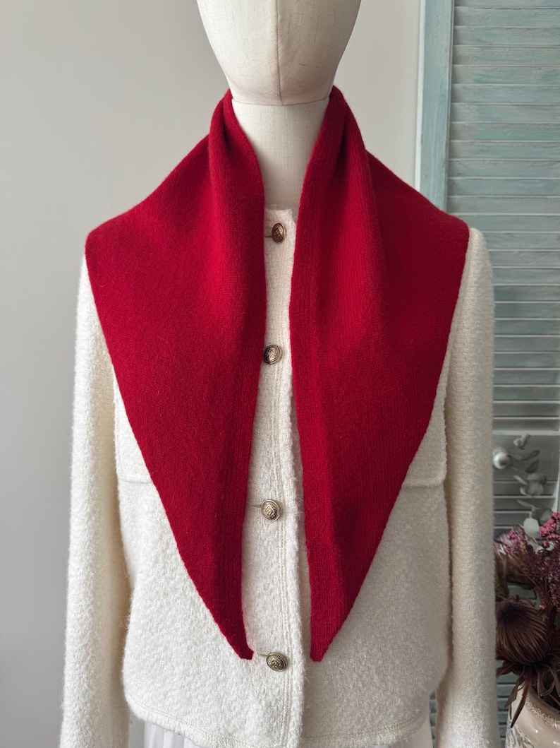 May include: A bright red triangular knit scarf draped over a cream-colored textured jacket. The jacket features a front button closure with three visible metallic buttons. The scarf has a smooth, solid-colored knit texture and pointed ends.