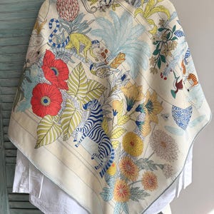 May include: A large square scarf featuring a cream background with a colorful print of a zebra, a monkey, birds, and various flowers including red poppies and yellow chrysanthemums. The scarf has a light blue border and is draped over a white shirt.