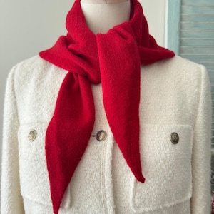 May include: A bright red triangular knit scarf tied around the neck, worn over a cream-colored textured jacket featuring two front pockets and four metallic buttons with a circular embossed design on the front placket.