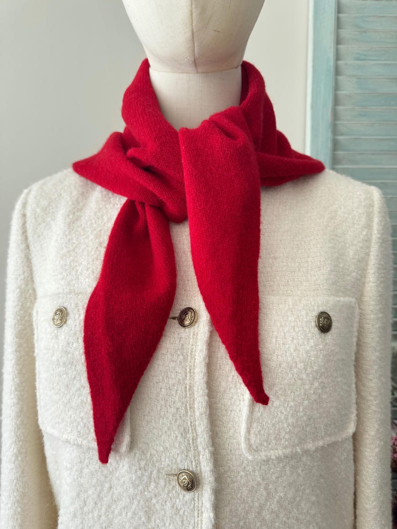 May include: A bright red triangular knit scarf tied around the neck, worn over a cream-colored textured jacket featuring two front pockets and four metallic buttons with a circular embossed design on the front placket.