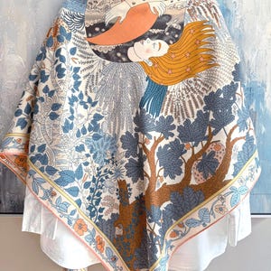 May include: A square scarf featuring a detailed illustration of a person with long orange hair resting against a large orange crescent moon. The design includes blue foliage, trees, and small flowers on a cream background with a decorative border.