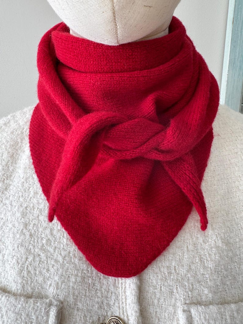 May include: A vibrant red triangular scarf with a knitted texture, wrapped and knotted around the neck of a mannequin. The scarf features a soft, fuzzy appearance and is draped to form a point at the front, layered over a white textured garment.