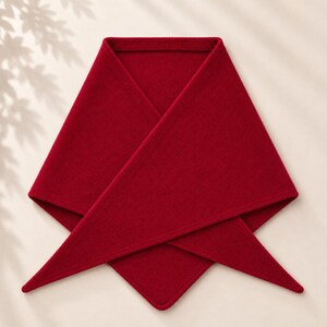 May include: A deep red triangular scarf featuring a fine knit texture. The garment is folded into a symmetrical shape with two long, pointed ends extending downwards and a wide, flat top edge, showcasing a consistent solid color throughout.