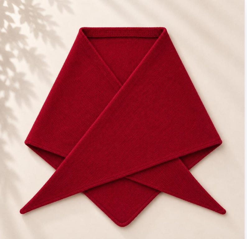 May include: A deep red triangular scarf featuring a fine knit texture. The garment is folded into a symmetrical shape with two long, pointed ends extending downwards and a wide, flat top edge, showcasing a consistent solid color throughout.
